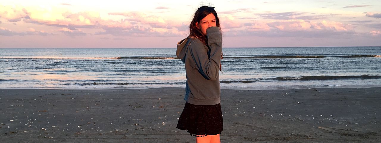 Portrait of young woman standing at beach against cloudy sky during sunset