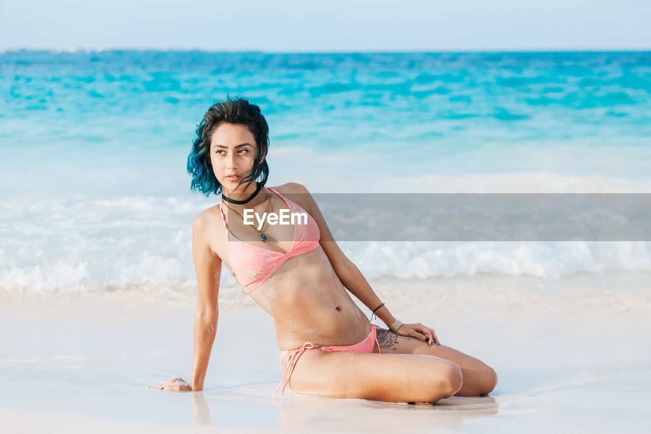 Woman in bikini kneeling on shore at beach against sky