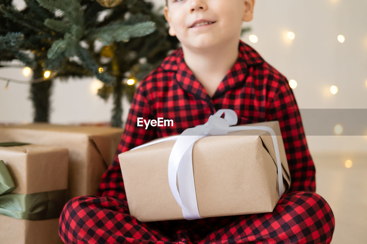 Cute little child boy in red pajama with gift boxes sitting under christmas tree at home