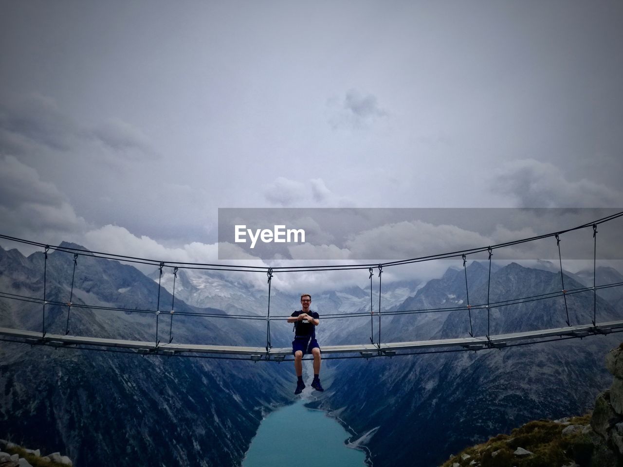 Man sitting on footbridge over mountains against sky