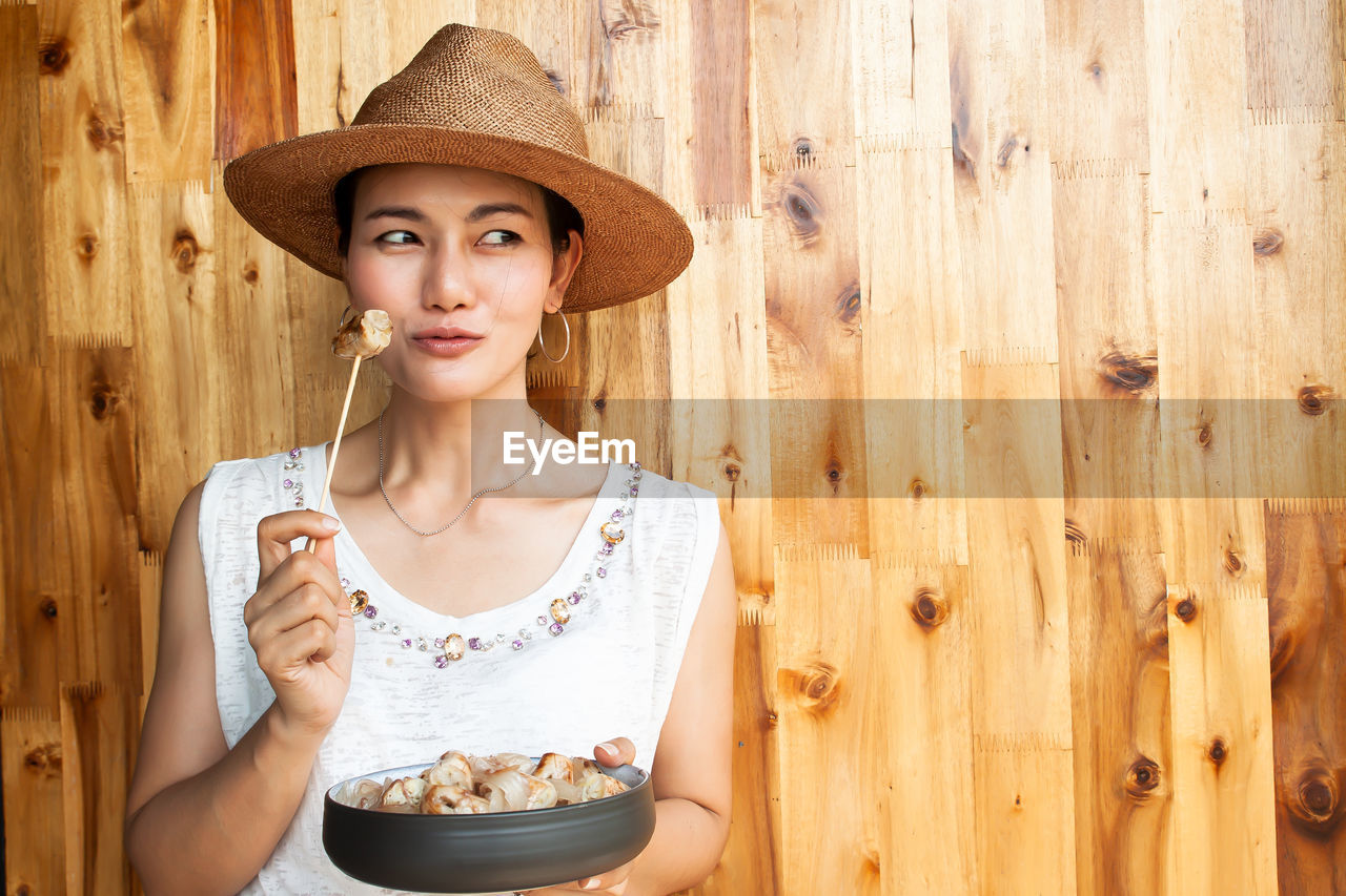 Thoughtful woman having food while standing against wooden wall