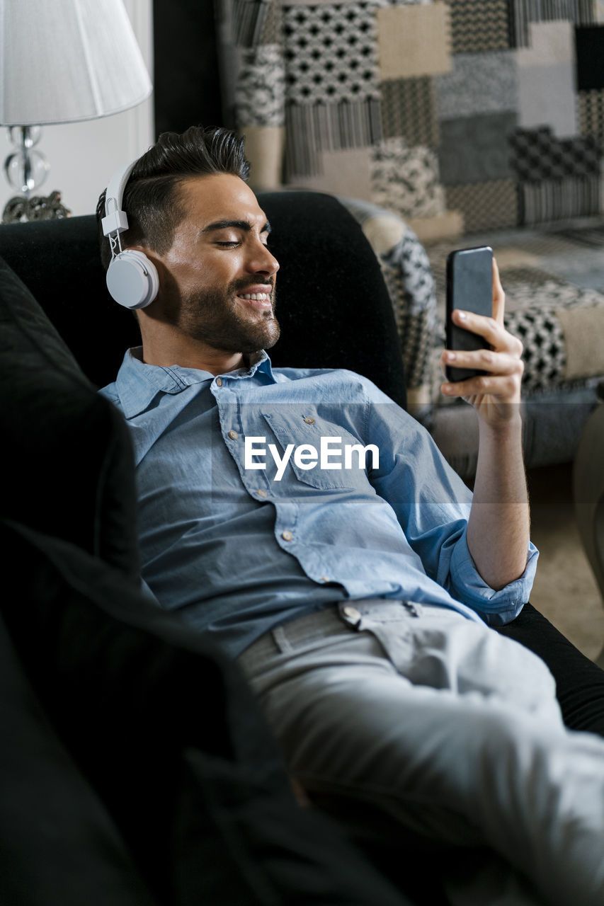 Smiling man wearing headphones using mobile phone while lying on sofa at home