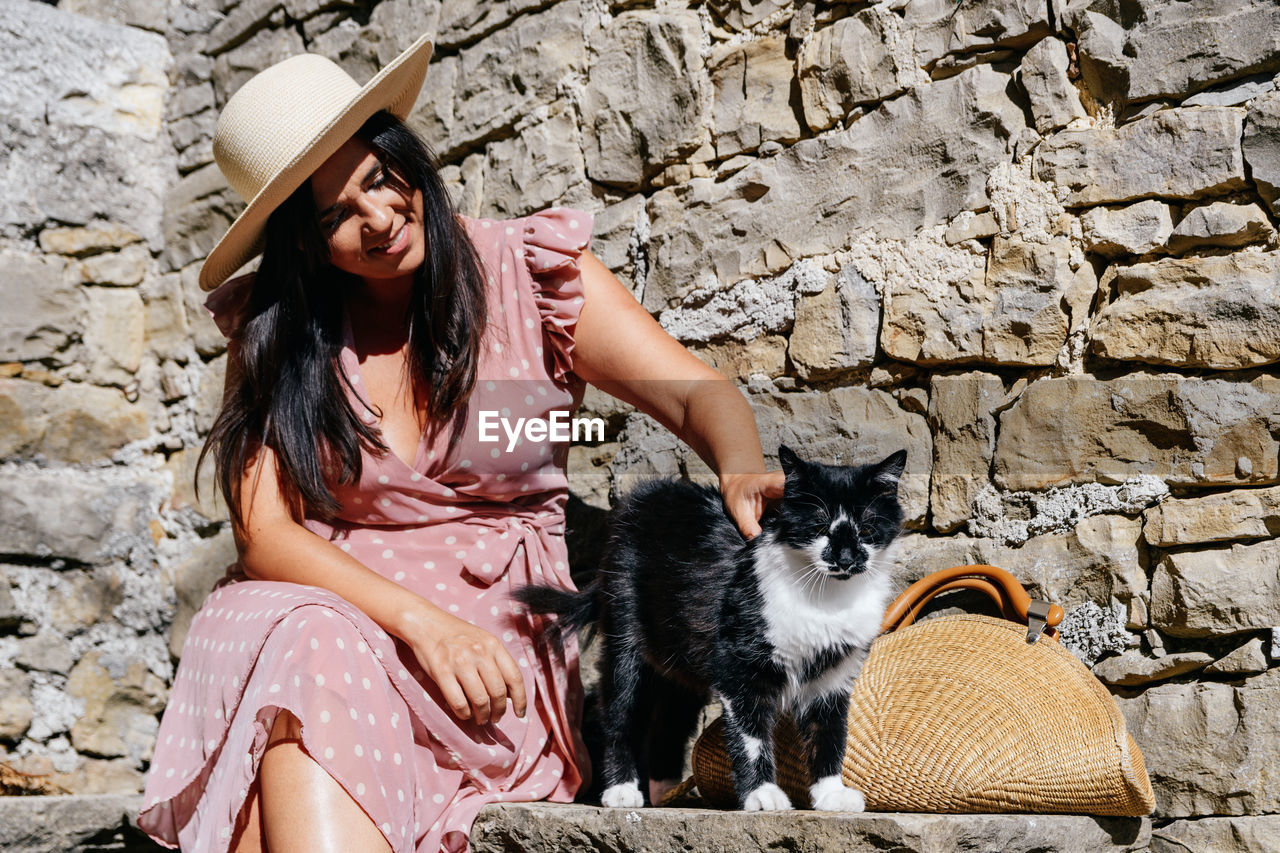 Young woman in pink dress sitting on stone bench in old town, petting adorable cat.