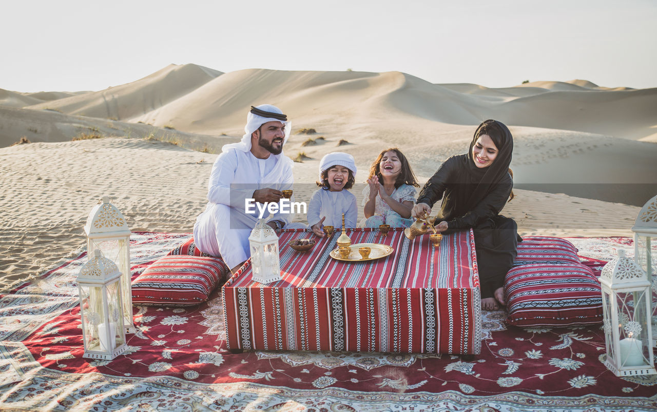 Arabian family having drink while sitting at desert against sky