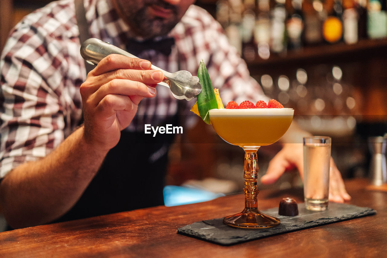 Unrecognizable barkeeper putting leaf on tropical cocktail with raspberries and pineapple near chocolate candy and liquor in pub