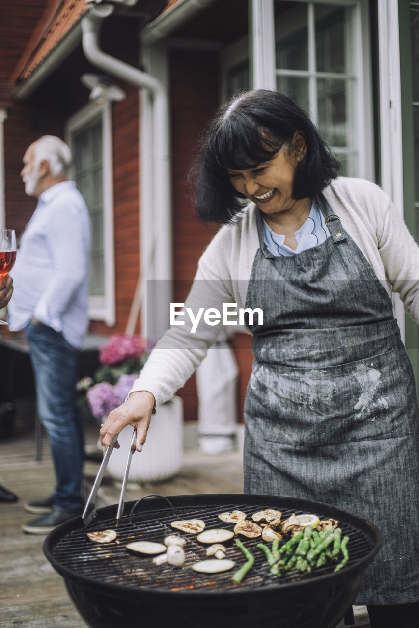 Happy woman wearing apron grilling vegetables on barbecue grill at party