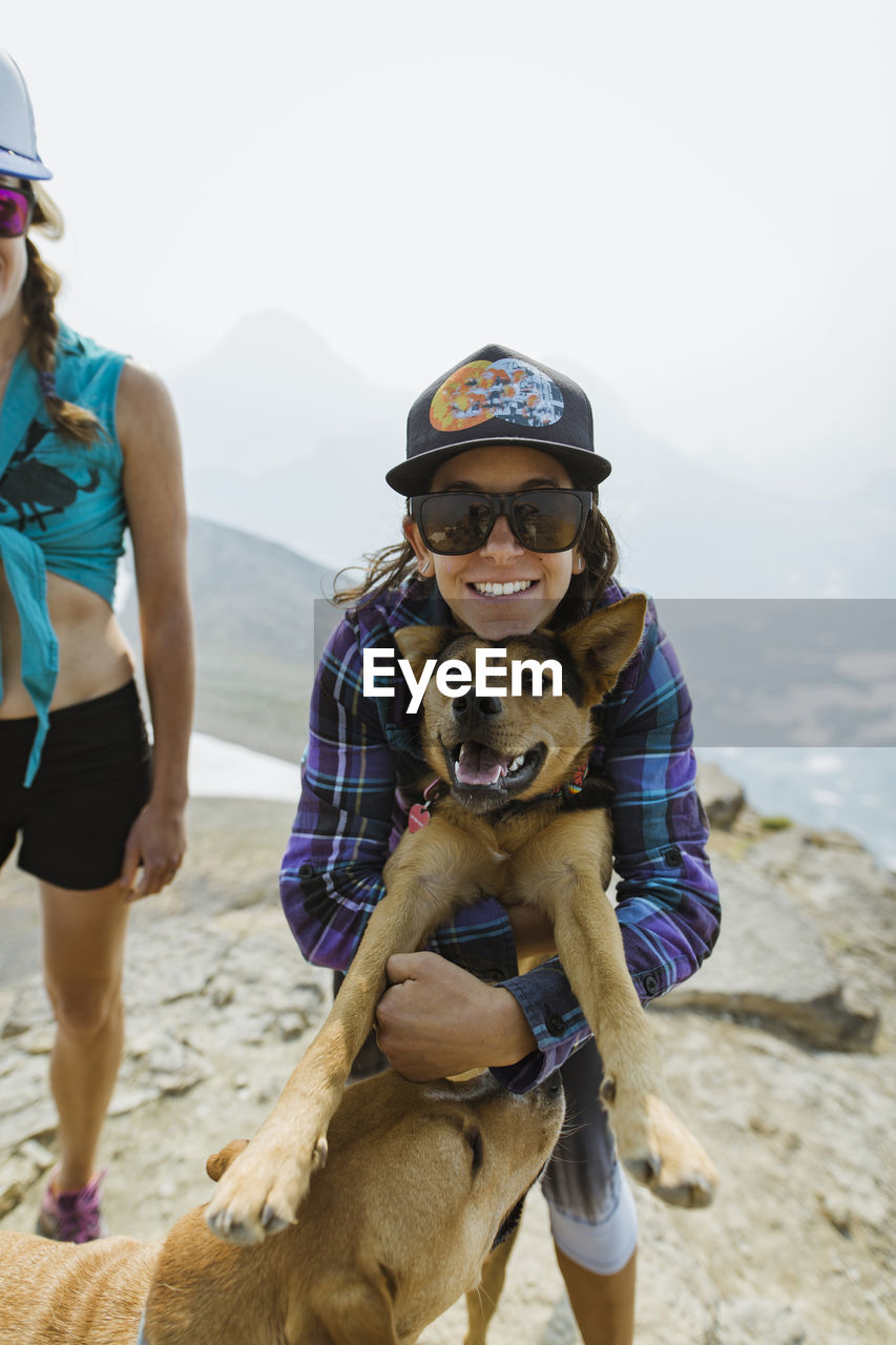 Portrait of woman embracing dog while standing by friend on mountain against clear sky