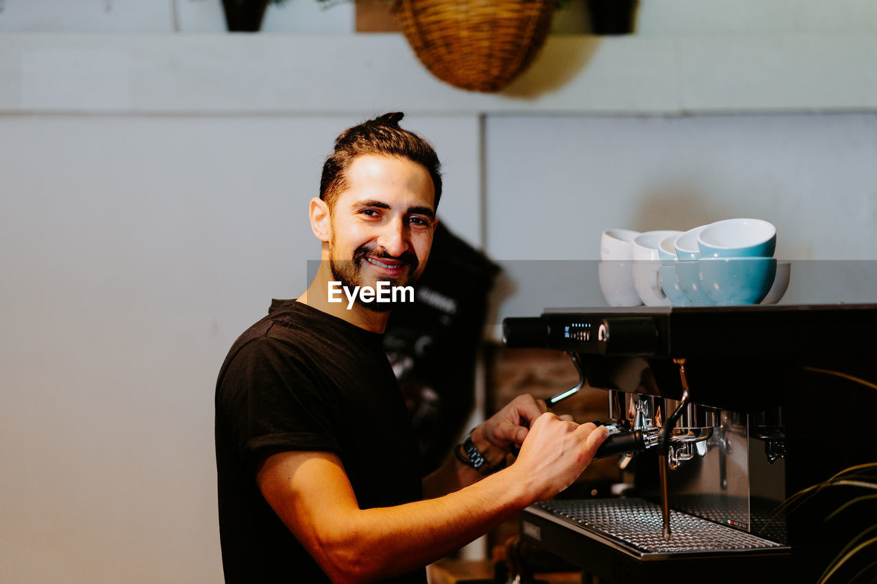 Side view of smiling male barista using portafilter and preparing coffee in modern coffeemaker while standing at counter in cafe and looking at camera