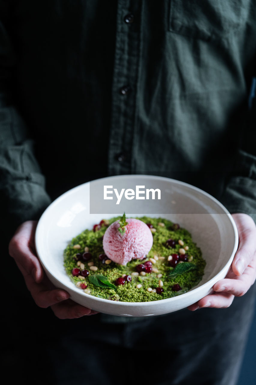Cropped person hands holding a white bowl with a scoop of purple ice cream on green mousse decorated with nuts and fresh mint