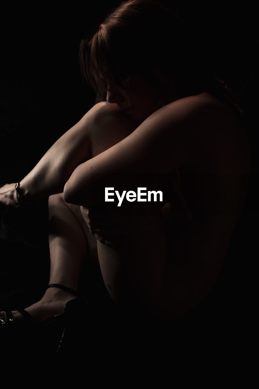 Naked woman hugging knees while sitting against black background