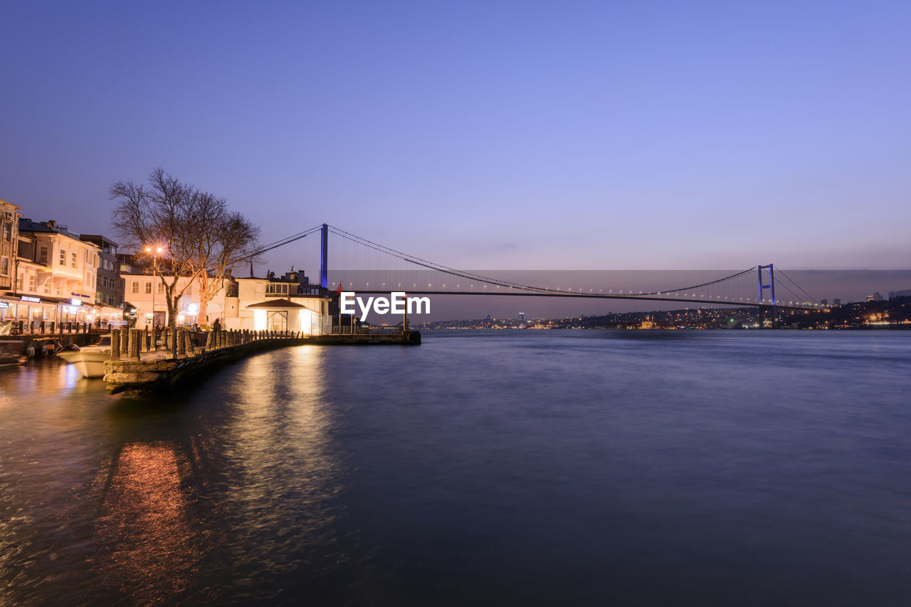 Illuminated suspension bridge over river against clear sky