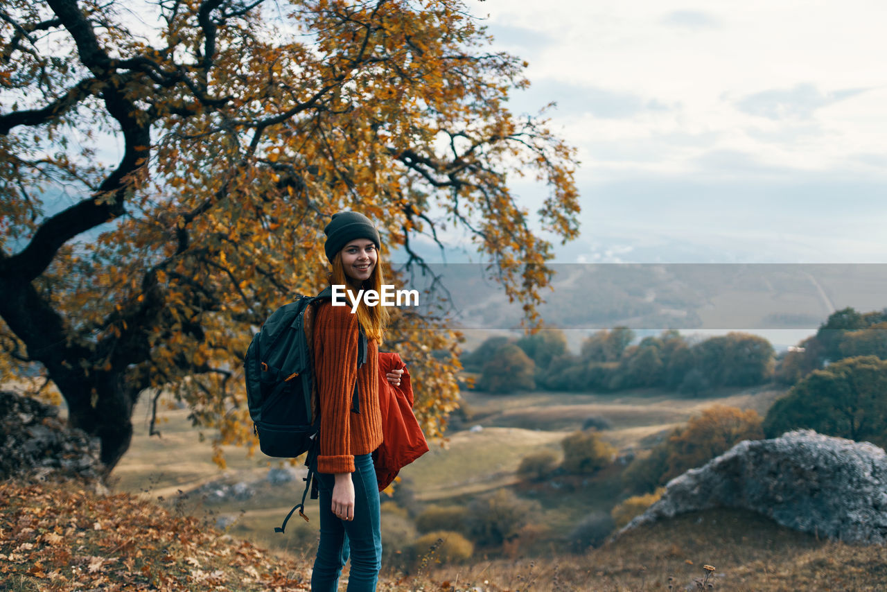 PORTRAIT OF YOUNG WOMAN STANDING BY TREE AGAINST PLANTS