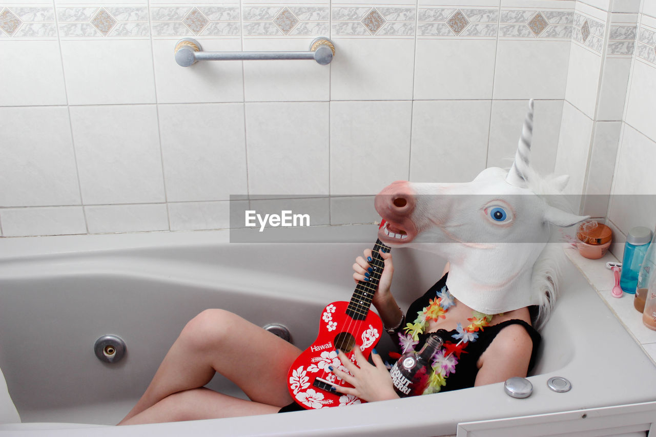 Woman wearing horse mask sitting with guitar in bathtub at home
