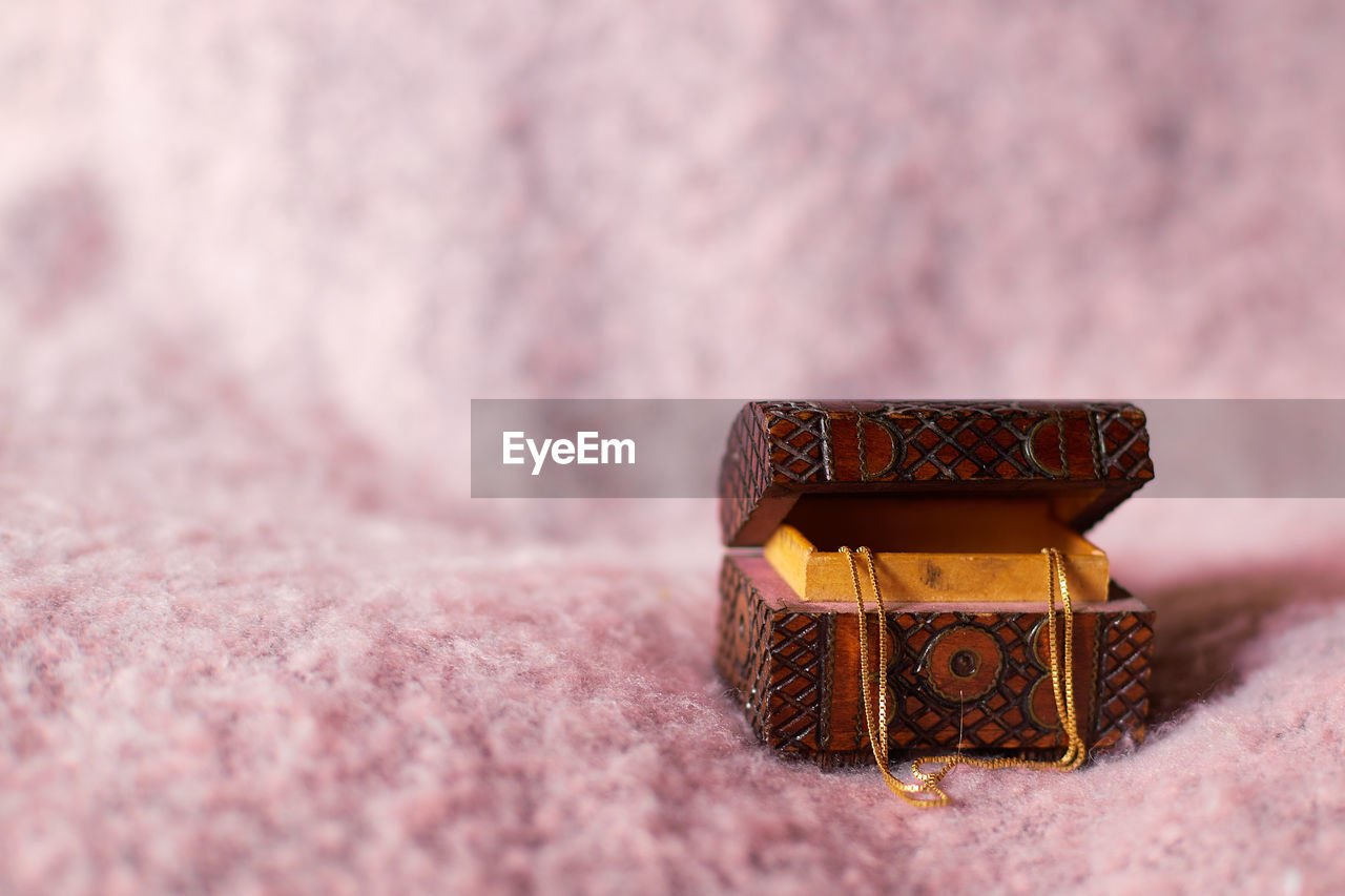 A tiny ornamented wooden jewelry box on pink background, with a golden chain hanging , offcenter