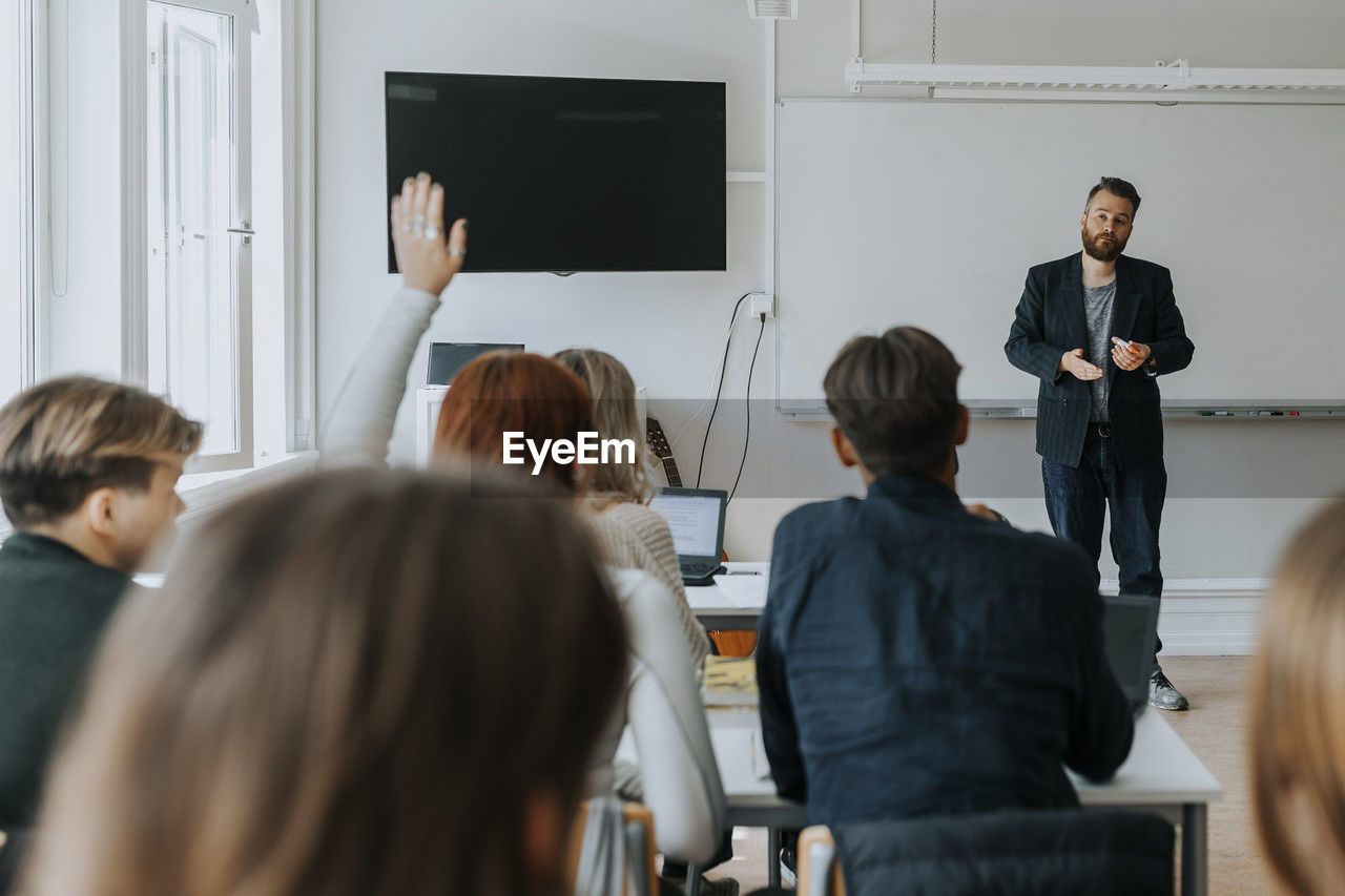 Male professor teaching while female student sitting with hand raised in classroom