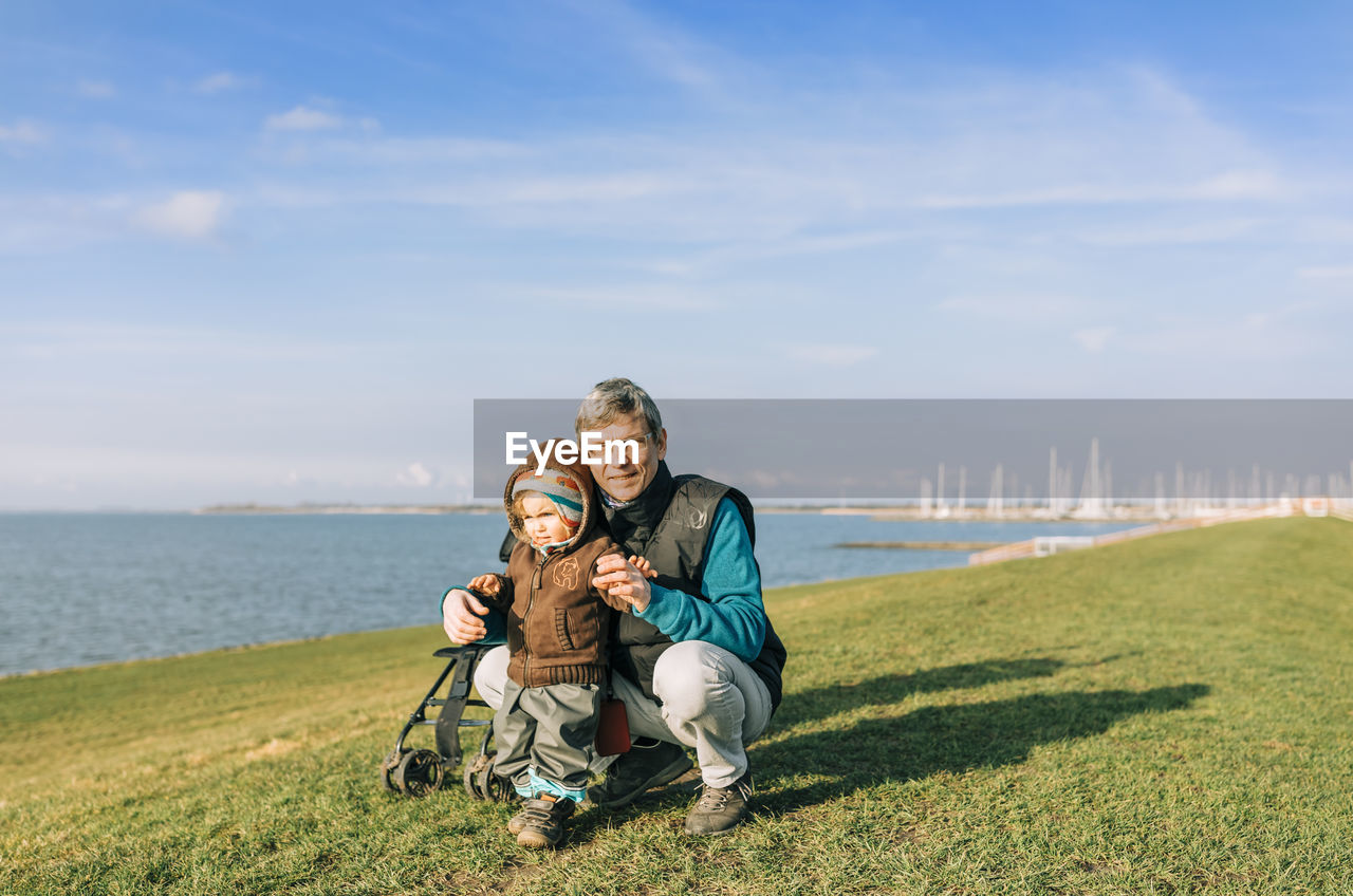 Grandfather holding granddaughter standing on grassy field against sea