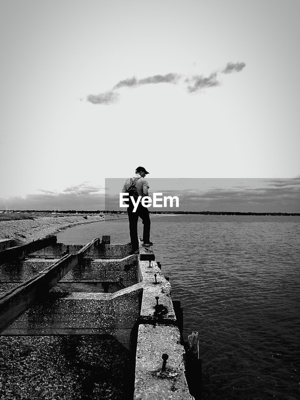 Rear view of man with backpack standing on retaining wall by sea against sky