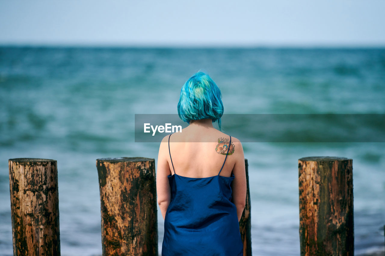 Beautiful blue-haired woman in long dark blue dress with cute tattoo on her shoulder blade on beach