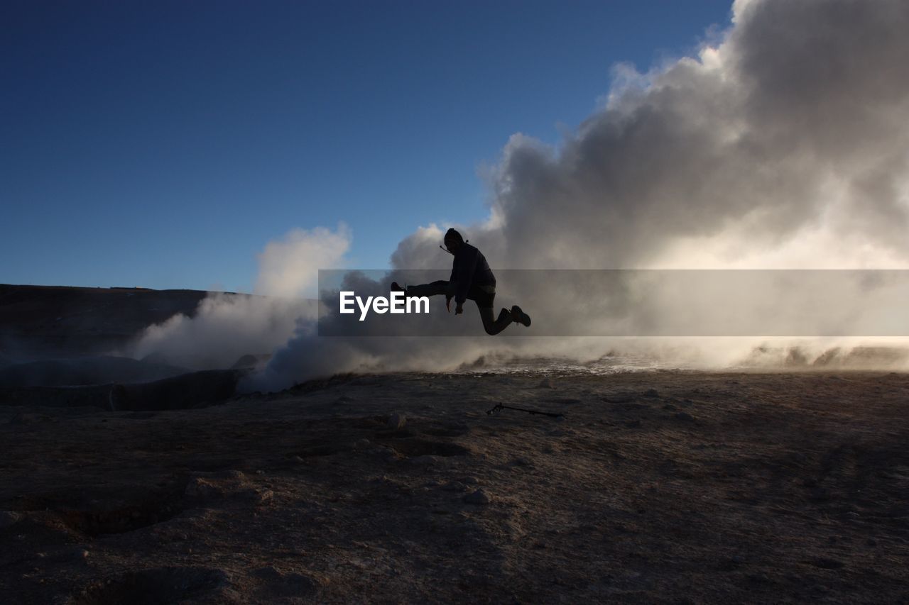 Silhouette man jumping by hot spring emitting steam against sky