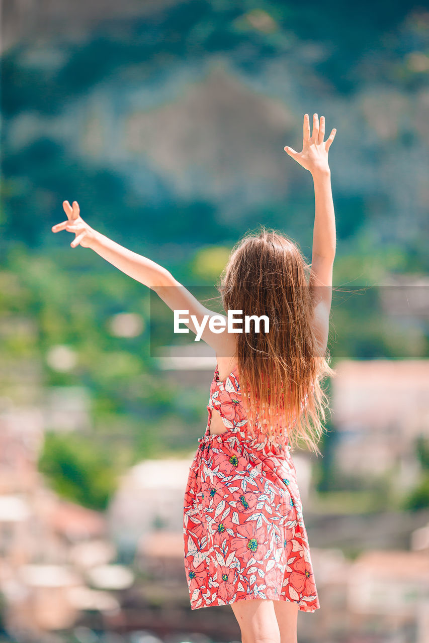 REAR VIEW OF WOMAN WITH ARMS RAISED STANDING IN PARK