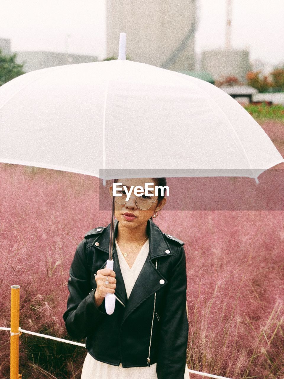 Portrait of young woman holding umbrella while standing against field in rain