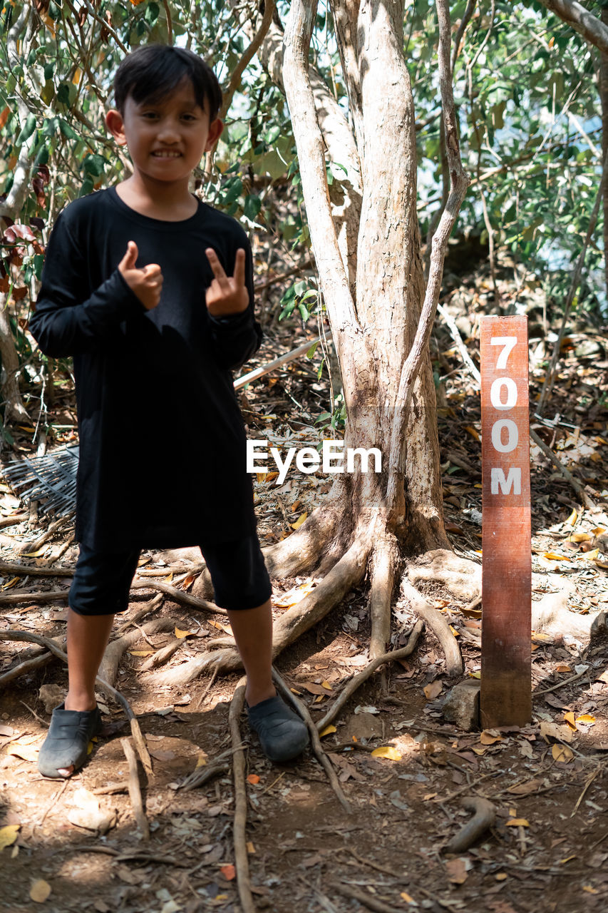 Full length portrait of boy gesturing while standing by tree trunk