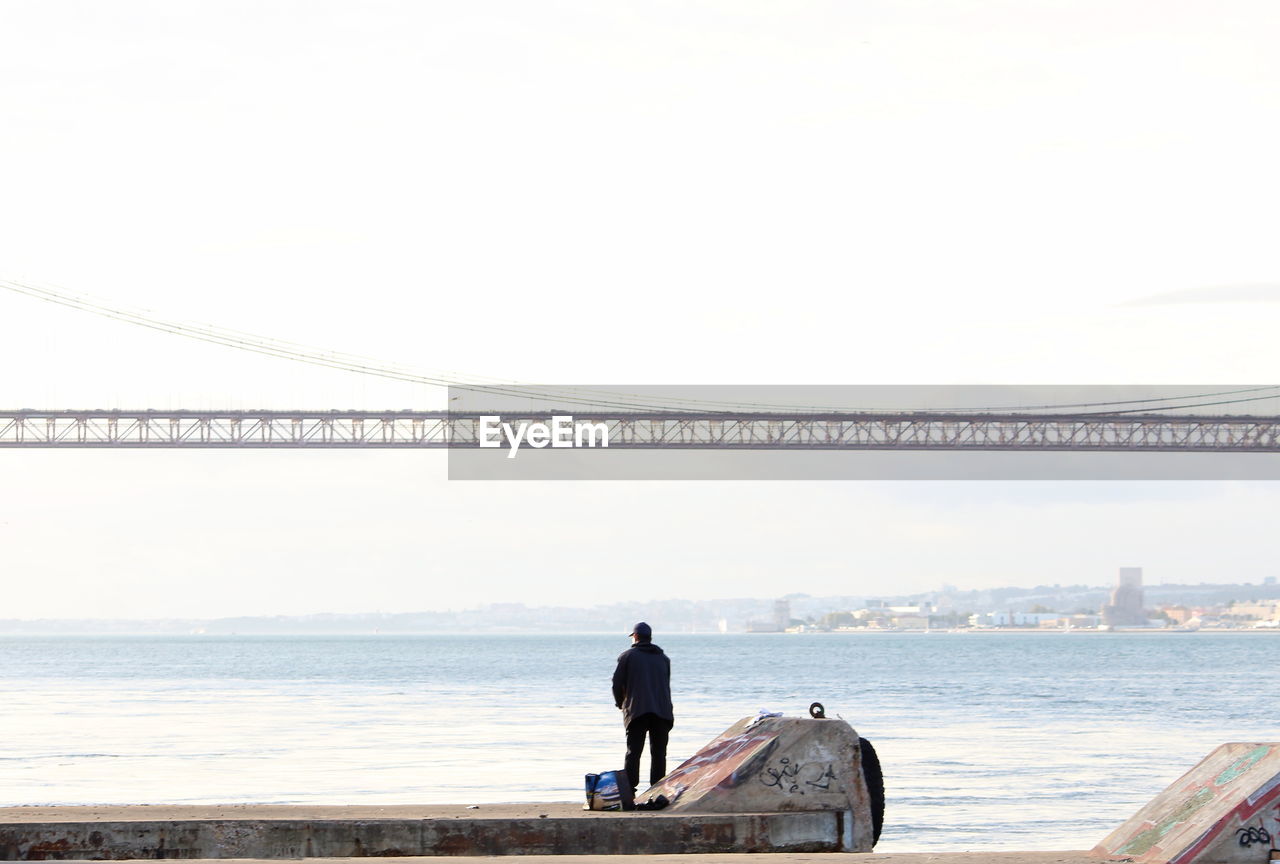 REAR VIEW OF MAN STANDING ON BRIDGE AGAINST SKY