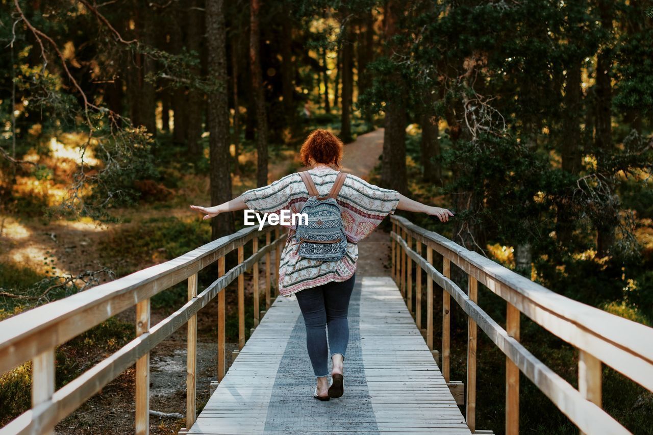 Rear view of woman with arms outstretched walking on footbridge in forest