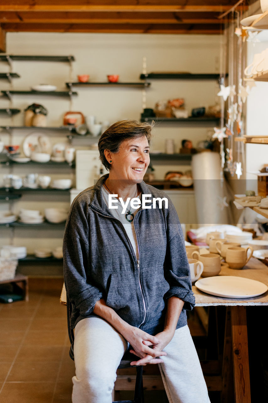 Happy middle aged female artisan looking away while resting after work in creative pottery studio