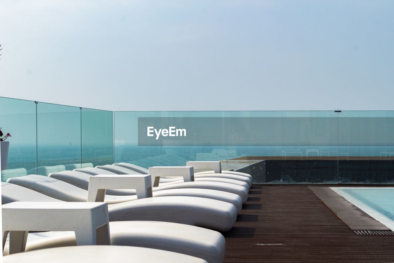 EMPTY CHAIRS BY SWIMMING POOL AGAINST SEA