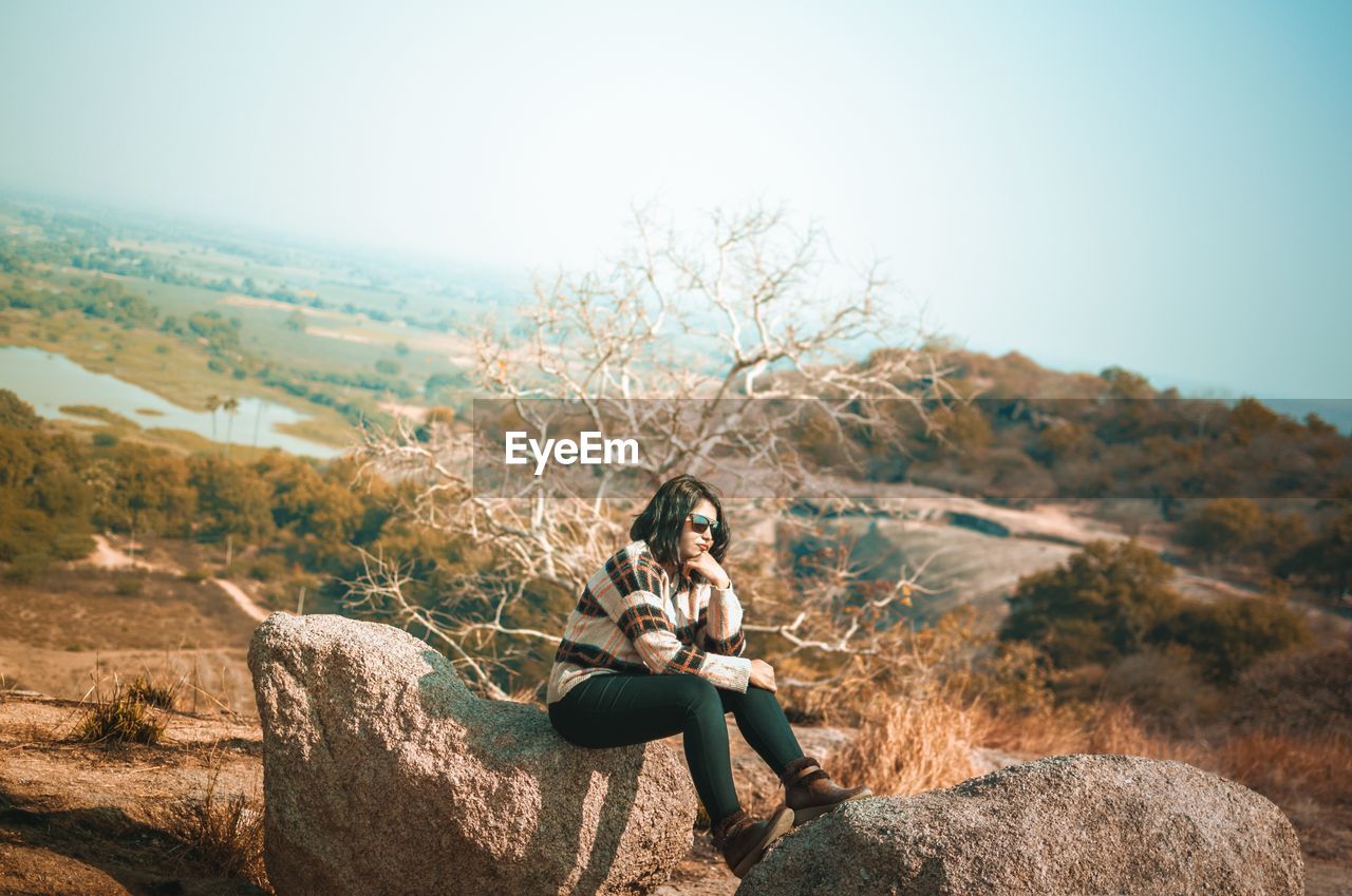 Woman wearing sunglasses sitting on rock against sky during sunny day