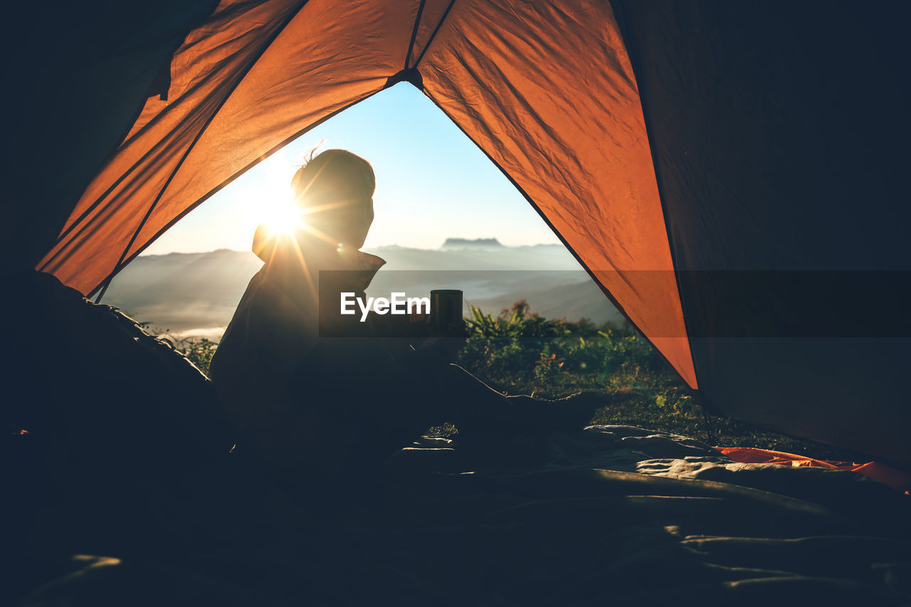 Silhouette man with mug in tent on mountain against bright sun in sky during sunrise