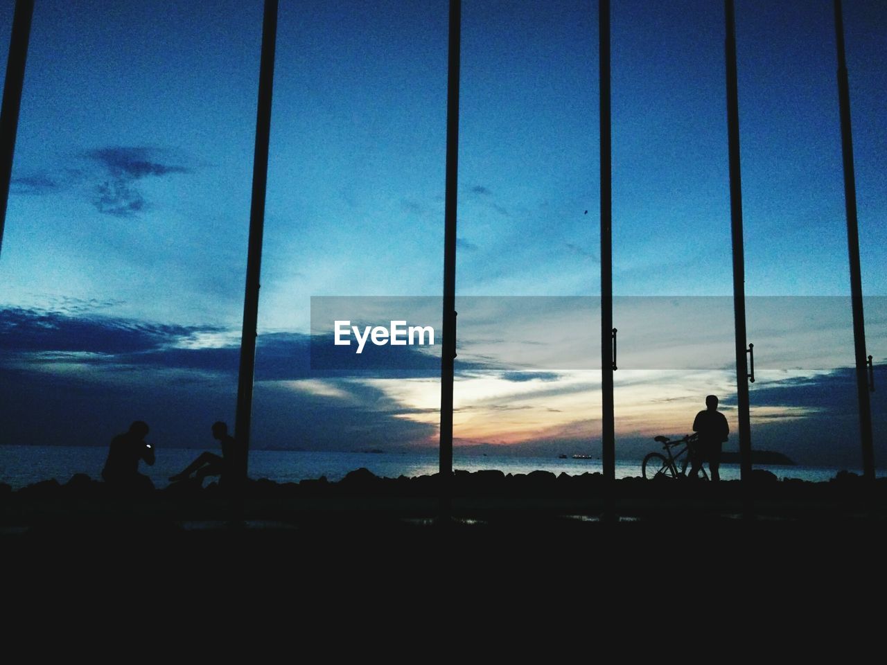 Silhouette people standing at beach against sky during sunset seen through gate