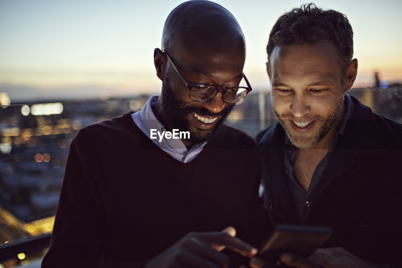 Smiling male professionals looking at mobile phone while standing on terrace during sunset