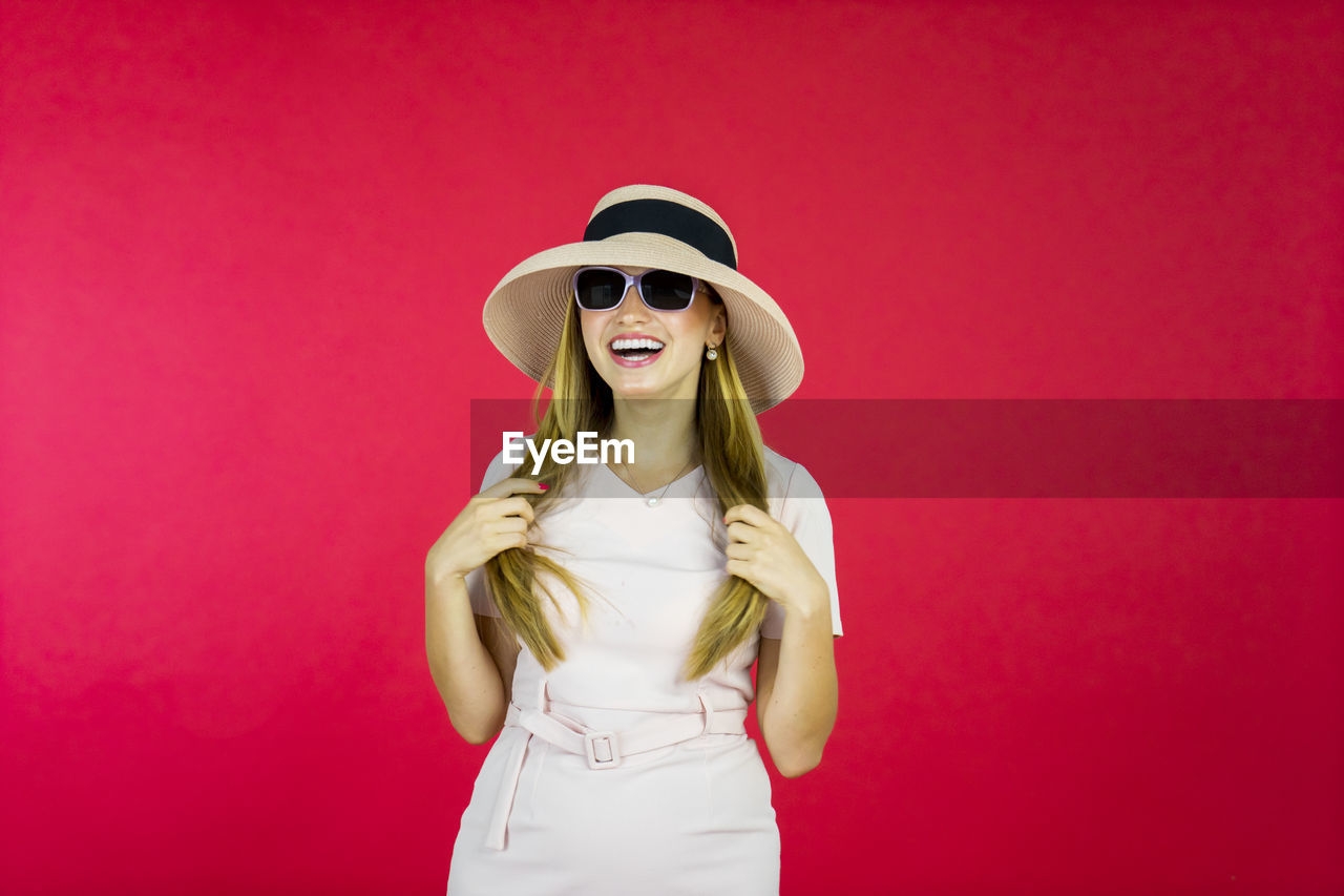 YOUNG WOMAN IN SUNGLASSES STANDING AGAINST RED BACKGROUND