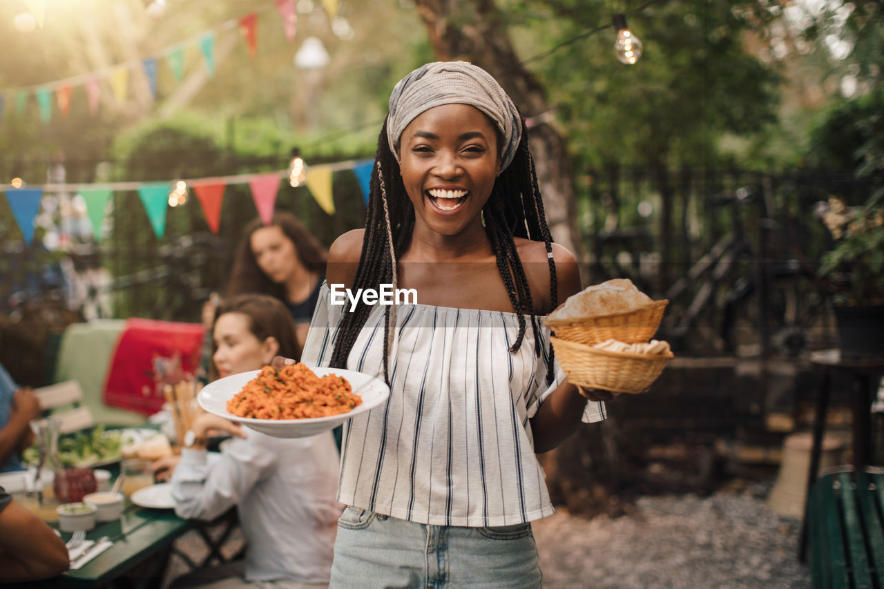 Portrait of smiling young woman carrying food while standing in backyard during garden party