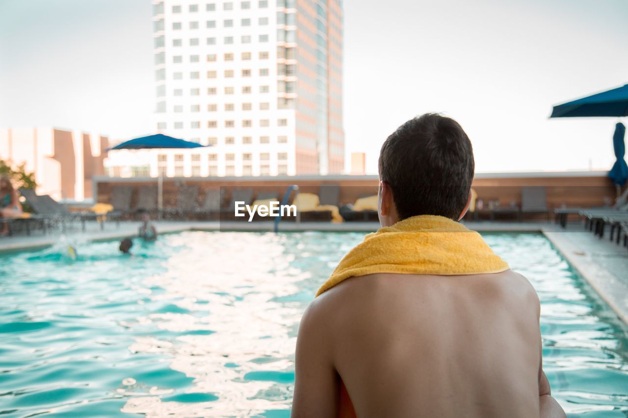 REAR VIEW OF SHIRTLESS MAN IN SWIMMING POOL BY BUILDINGS