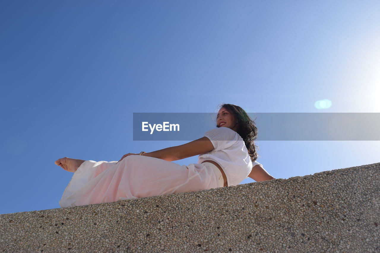 Low angle view of woman sitting on retaining wall against clear blue sky