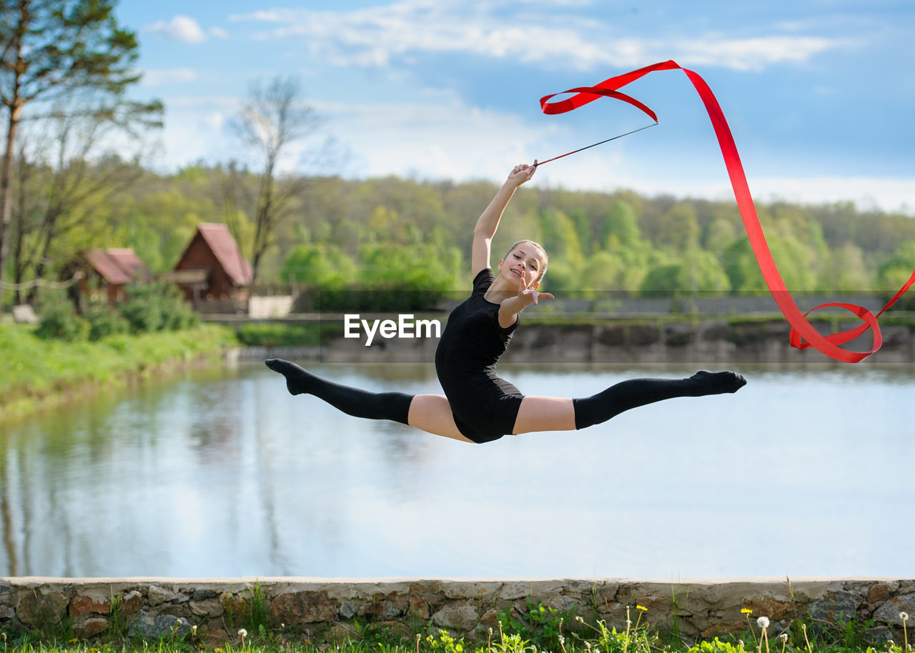 Full length portrait of woman gymnast jumping in air while holding ribbon against lake