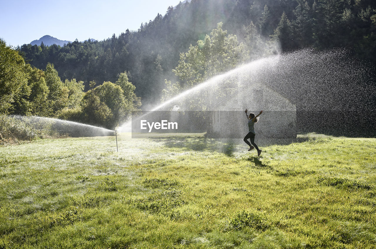 Woman jumping on grass near sprinkler during sunny day