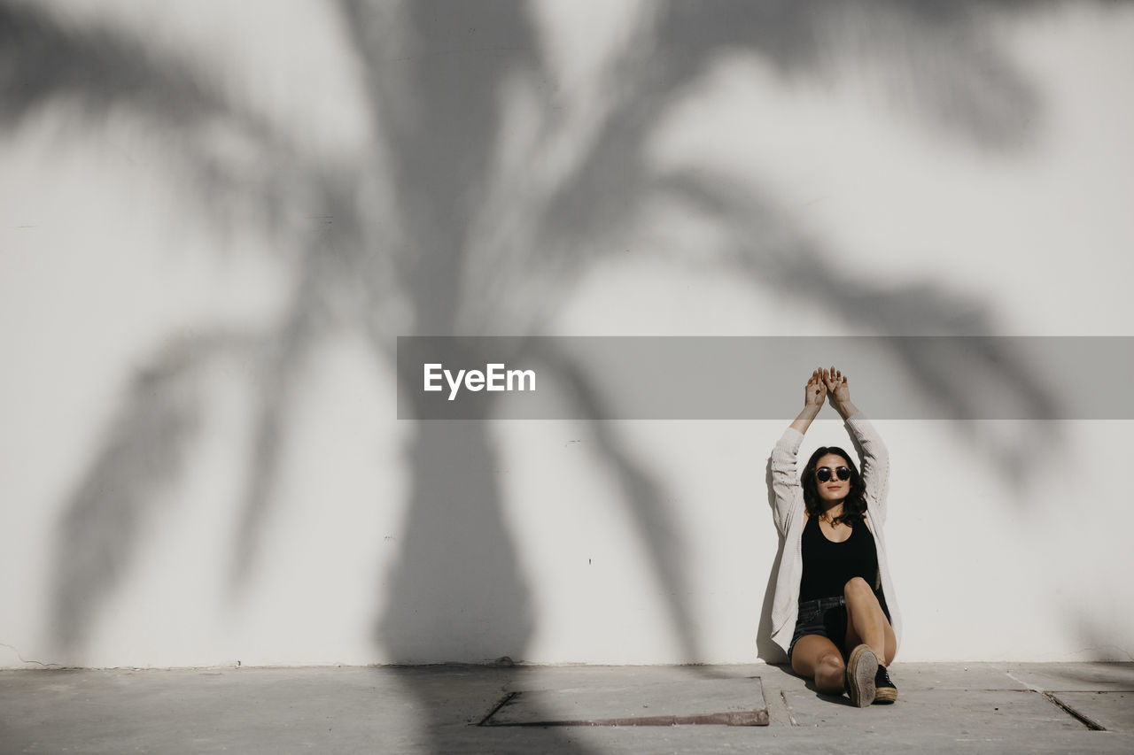 Young woman with arms raised sitting by palm tree shadow on white wall