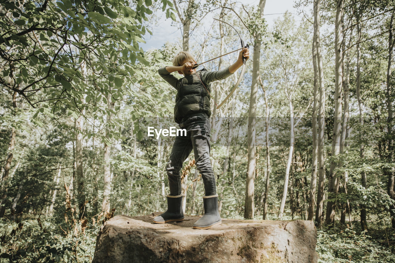 Boy aiming with bow and arrow while standing on boulder in forest