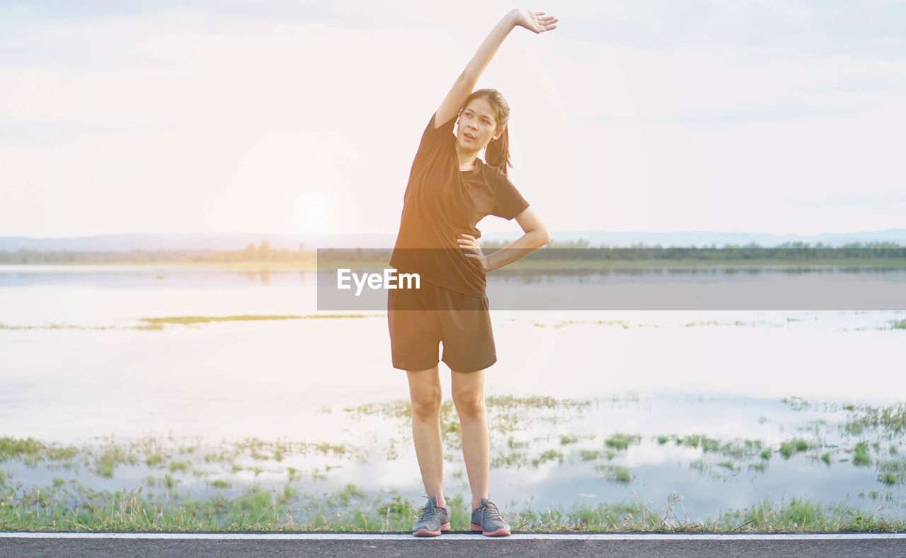 Young woman stretching on road against lake and cloudy sky