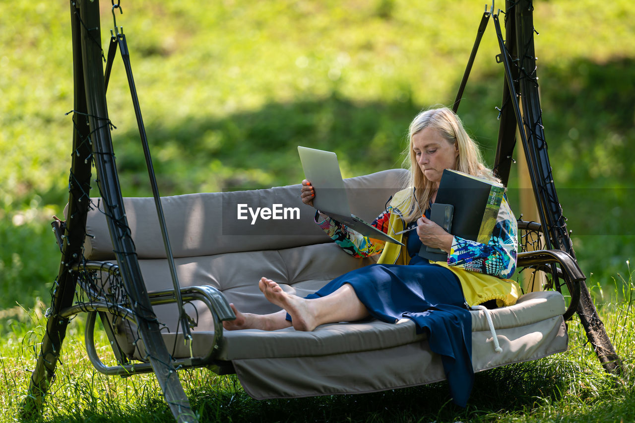 Mature woman with laptop and documents working in garden on rocking couch, green home office concept