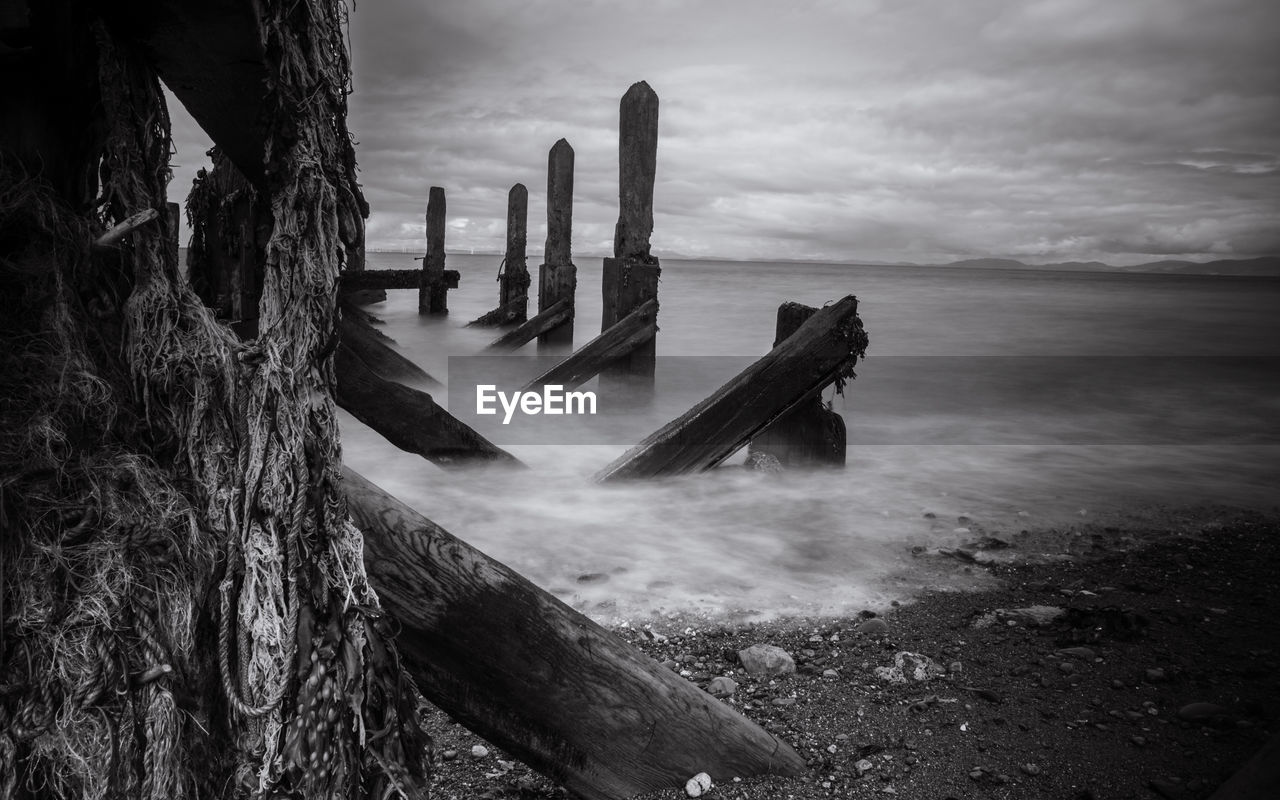 Abandoned wooden structure at beach against cloudy sky