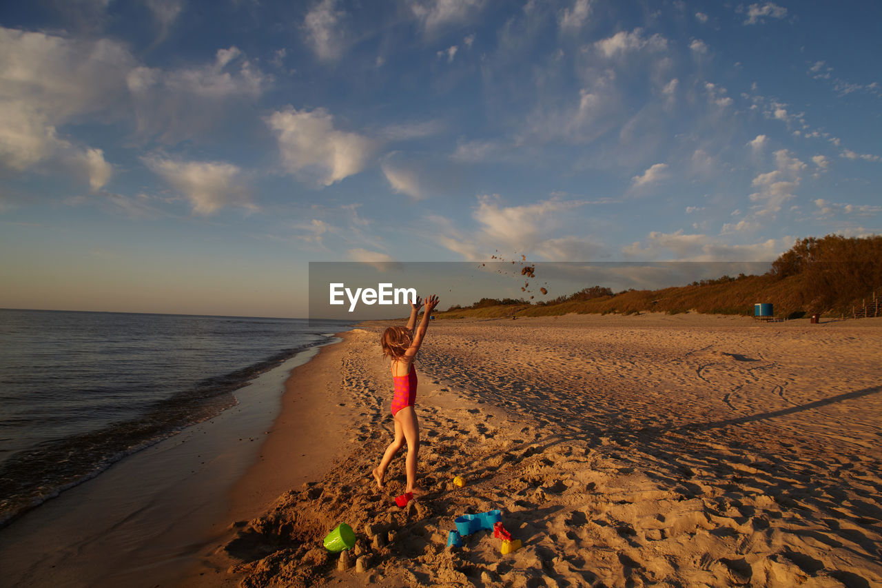 Girl with arms raised standing at beach against sky during sunset