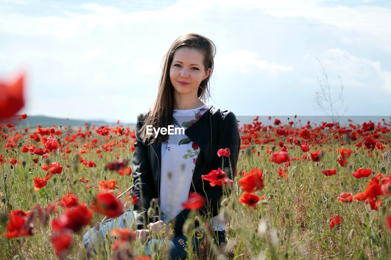 Portrait of young woman standing by poppy flowers on field against sky