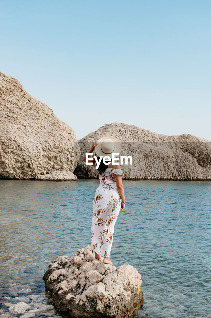 Rear view image of woman in long summer dress standing on rock on beautiful beach.