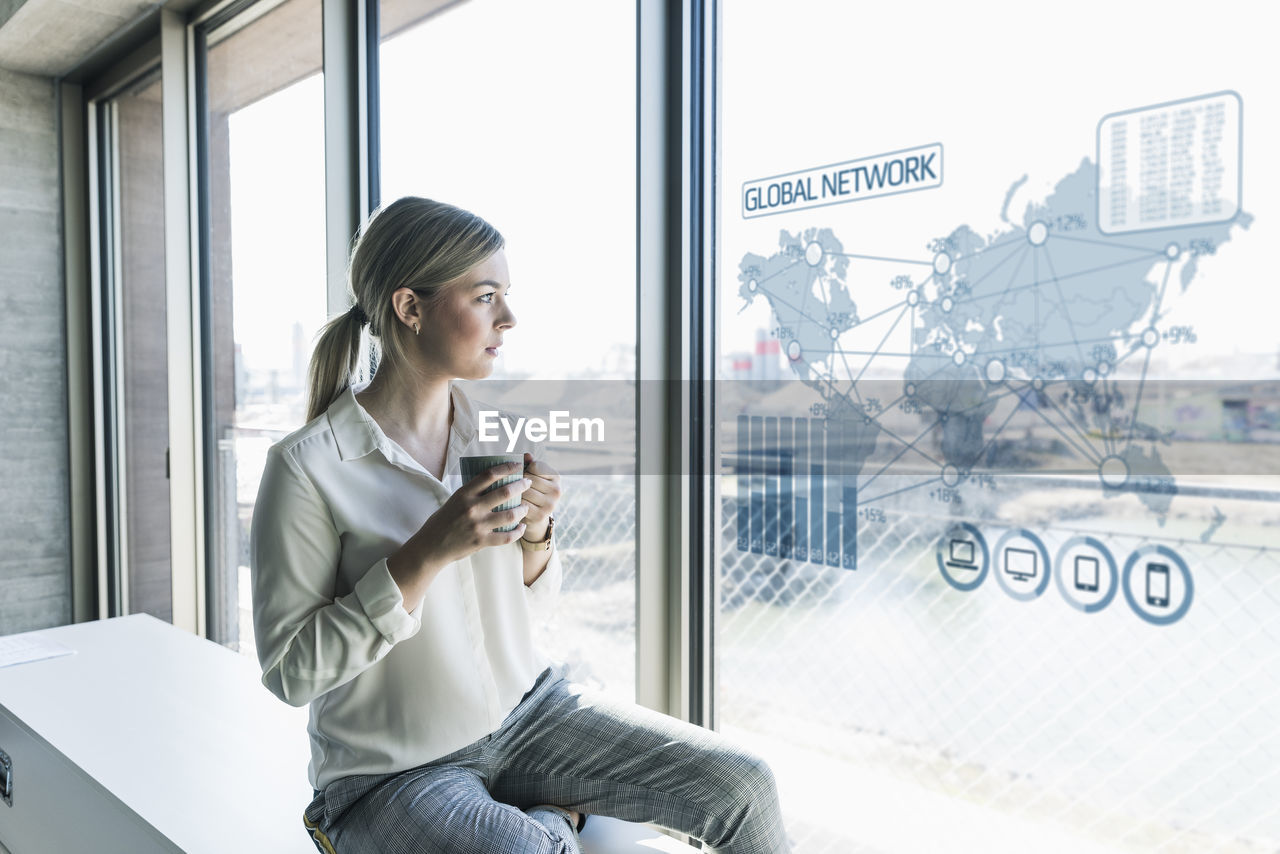 Young businesswoman looking at virtual world map at window pane in office