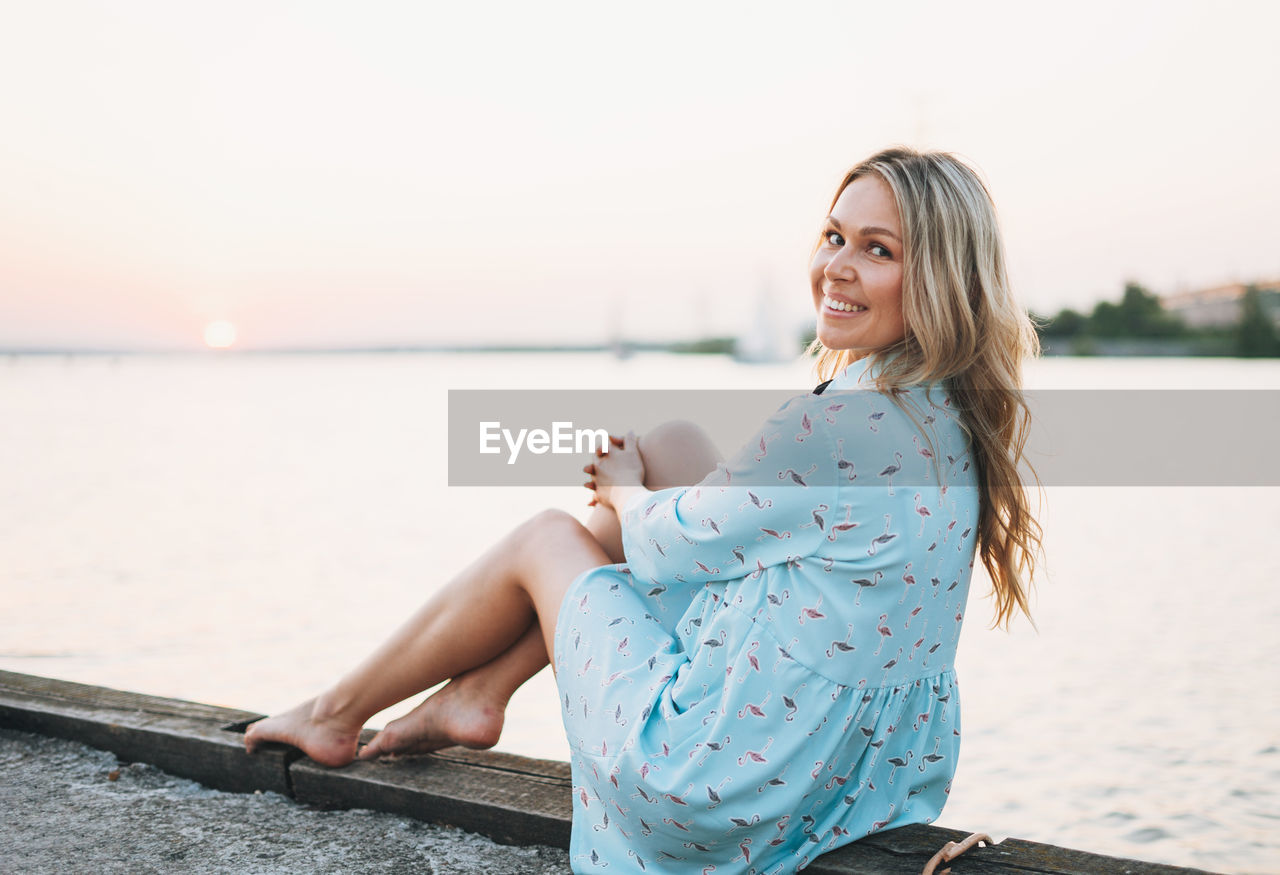 Beautiful blonde young woman in blue dress sitting on pier and looking on sunset